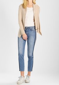 Beige ribbad cardigan yliä vaalean yläosan, yhdistettynä sinisiin farkkuihin, joissa on mustat sivusuunnat, ja valkoisiin nauhakengiin.