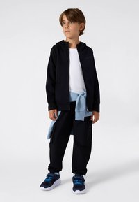 Svart hettejakke med glidelås, hvit T-skjorte, løse svarte cargobukser, lyseblå genser knyttet i livet, og mørke joggesko med blå detaljer.