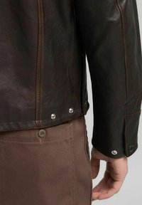 Giacca in pelle marrone con texture liscia, caratterizzata da cuciture a contrasto, bottoni a pressione e design aderente. Pantaloni marroni visibili.