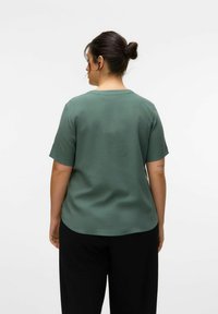 Top de manga corta verde de tela suave con un cuello redondeado y un dobladillo curvado, acompañado de pantalones negros holgados.