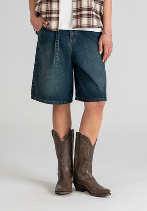 Shorts en jean bleu foncé à coupe ample, associés à des bottes de cowboy en cuir marron ornées de broderies et à bouts pointus.