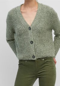 Hellgrüner gestrickter Cardigan mit V-Ausschnitt, fünf dunklen Knöpfen vorne und langen Ärmeln; kombiniert mit dunkelgrünen, eng anliegenden Hosen.