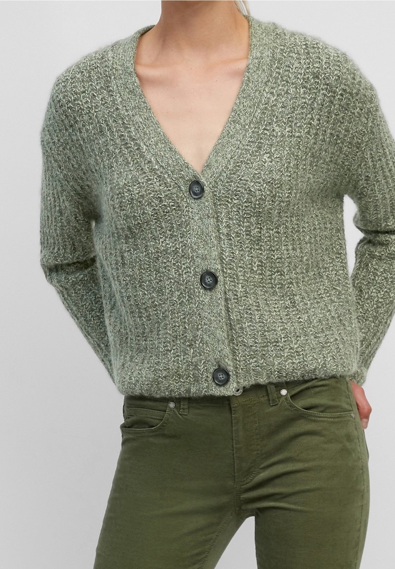 Hellgrüner gestrickter Cardigan mit V-Ausschnitt, fünf dunklen Knöpfen vorne und langen Ärmeln; kombiniert mit dunkelgrünen, eng anliegenden Hosen.