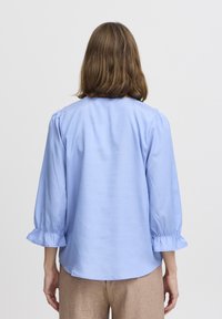 Camicia azzurro chiaro con maniche a puff e polsini arricciati, caratterizzata da una texture liscia e una vestibilità comoda, abbinata a pantaloni in lino color sabbia.