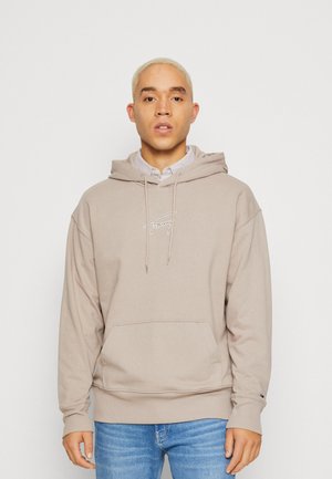 Felpa beige con cappuccio, tasca frontale, coulisse e logo ricamato. Tessuto morbido con vestibilità rilassata, abbinata a jeans azzurri.