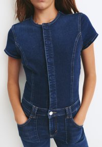 Dżinsowy kombinezon z krótkim rękawem, w ciemnoniebieskim kolorze, z zapięciem na guziki z przodu, dwiema przednimi kieszeniami i kontrastowym przeszyciem wzdłuż szwów.