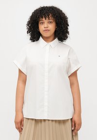 Chemise blanche à manches courtes avec un col, ornée d'un petit logo coloré sur la poitrine et d'une texture de tissu lisse et légère.
