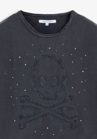 Camiseta negra de algodón con diseño de calavera y tibias cruzadas, adornada con pequeños remaches plateados. Bordes sin rematar y cuello redondo.