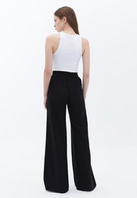 Pantaloni neri a gamba larga realizzati in un tessuto morbido, con una vita elasticizzata, abbinati a un top bianco aderente, visti da dietro su uno sfondo bianco.