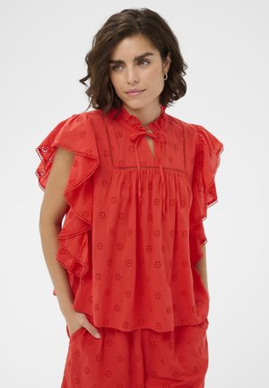 Kobieta o krótkich ciemnych włosach, ubrana w czerwoną bluzkę z ażurowym wzorem i falbankami na rękawach oraz czerwone krótkie spodenki, ręce w kieszeniach, patrzy nieco w prawo.