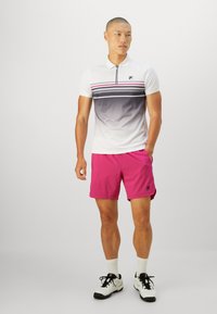 Man standing with hands in pockets wearing white and gray gradient polo, bright pink shorts, white socks, and white athletic shoes.