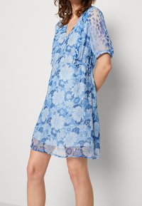Robe florale bleue avec un décolleté en V, des manches bouffantes et un voile transparent à la texture douce et aux motifs floraux superposés.