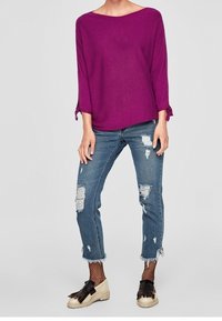 Haut violet à manches trois-quarts avec un décolleté bateau. Porté avec un jean bleu clair déchiré, présentant un ourlet effiloché et des chaussettes en résille dans les chaussures.
