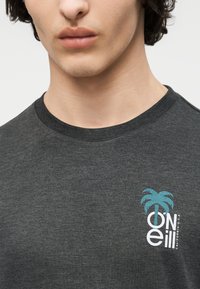 Ung person som har på seg en mørkegrå t-skjorte med rund hals og et turkist palmetre samt hvit "O'Neill California USA"-logo på venstre bryst.