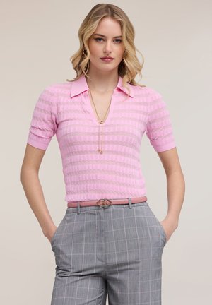 Mujer rubia con camiseta polo de manga corta rosa texturizada, pantalones a cuadros grises con cinturón rosa, de pie con las manos en los bolsillos.