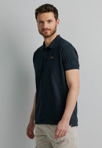 Marineblaue Polohemden mit kurzen Ärmeln, einem Kragen und einem kleinen Emblem auf der linken Brust. Baumwollstoff mit glatter Textur.