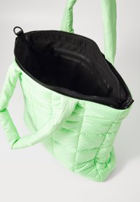 Green puffy tote bag with quilted texture, zippered black interior, and adjustable straps. Features a hidden pocket and smooth material.