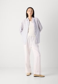 Chemise boutonnée rayée bleu et blanc, débardeur blanc, pantalon en lin blanc et ballerines beiges. Texture lisse, coupe décontractée, couleurs claires.