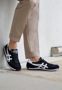 Chaussures de sport noir et blanc avec une tige en mesh et en daim. Elles présentent un embout arrondi et des bandes blanches sur le côté. Portées avec un pantalon beige clair.