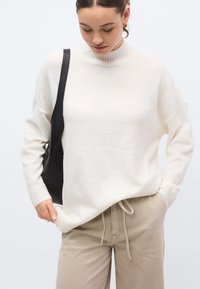 Kreminės spalvos megztinis su aukštu apykakliu ir juostelėmis rankogaliais, derinamas su smėlio spalvos kelnėmis su elastiniu liemeniu ir juoda petneša.