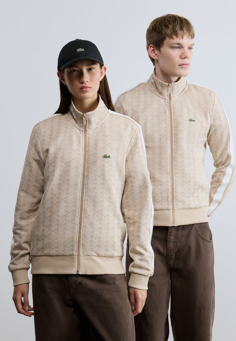 Μπεζ ζακέτες με φερμουάρ, με υφή γεωμετρικού μοτίβου, λευκές πλαϊνές ρίγες και πράσινο λογότυπο στο στήθος, συνδυασμένες με σκούρο παντελόνι και μαύρο καπέλο.
