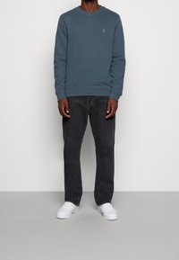 Niebieska bawełniana bluza z okrągłym dekoltem i ściągaczami na rękawach, w połączeniu z ciemnoszarymi jeansami i białymi trampkami, stojąca na jednolitym tle.