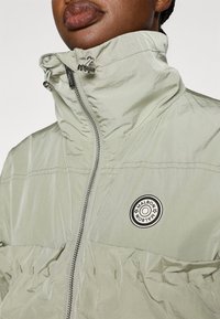 Ανοιχτό πράσινο τζάκετ με φερμουάρ, υψηλός γιακάς, κορδόνι και στρογγυλή μαύρη ετικέτα με λογότυπο στο στήθος, κατασκευασμένο από υφή υφάσματος.