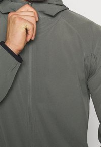 Chaqueta gris ligera con cremallera, capucha y puños elásticos. Cuenta con una textura suave y detalles de malla bajo las axilas.