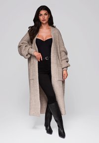 Strick-Langcardigan in hellem Beige, mit lockerer Passform, offener Vorderseite und gerippten Bündchen. Getragen über einem schwarzen Body und kombiniert mit schwarzen Hosen und Stiefeln.