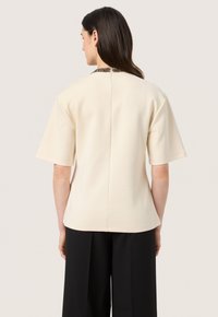 Beige kortærmet top med rund halsudskæring, tekstureret krave og lige snit. Glat stof med en detalje ved bagsiden.