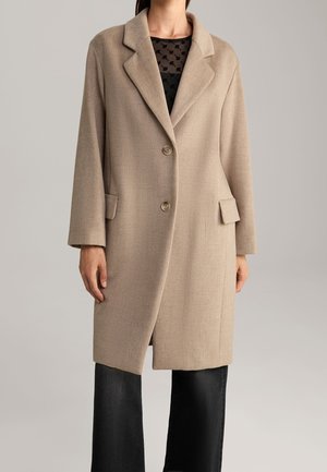 Kobieta ubrana w beżowy płaszcz do kolan z klapami, dwoma guzikami, bocznymi kieszeniami z patkami oraz czarne szerokie spodnie na tle o neutralnym kolorze.