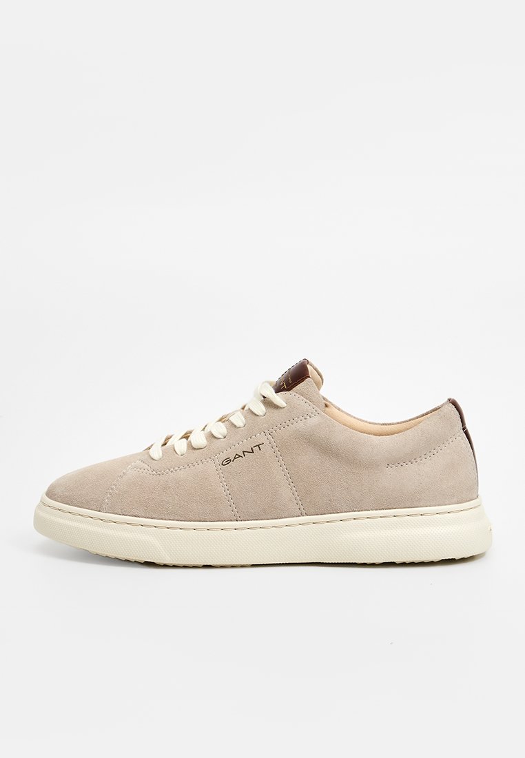 Beige mockask sneaker med rund tå, ton-i-ton sömnad, krämfärgade snörning och en brunt läderdetalj vid hälen. Varumärkeslogotyp på sidan.