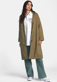 Abrigo largo de cuadro jefes marrón con un ajuste relajado, que presenta grandes botones, bolsillos laterales y una tela texturizada. Combinado con una camisa blanca y pantalones verdes.
