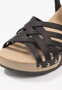 Zwarte leren sandaal met gekruiste bandjes, een natuurlijke houten basis en metalen accenten. Gestructureerde zool voor grip en ondersteuning.