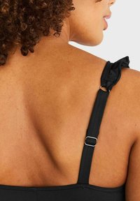 Svart simtop med justerbar rem som har en volangdetalj och metallhardware. Slät, texturerad material mot huden.