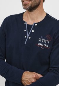 Tamsiai mėlynas ilgomis rankovėmis marškinis, pagamintas iš medvilnės, su henley tipo apykakle ir užsegamomis sagomis, dekoruotas baltu ir raudonu tekstu.