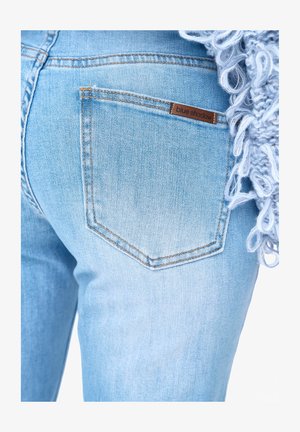 Tylnia kieszeń z jasnoniebieskiego denimu z brązową etykietą z napisem „blue shadow”, obok blado szarej dzianiny z pętlami.