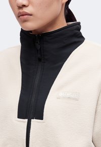 Κοντινό πλάνο ατόμου που φοράει μπουφάν fleece σε κρεμ και μαύρο χρώμα με ψηλό γιακά και εμφανές το λογότυπο της Columbia Sportswear στο στήθος.