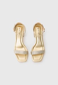 Zlate kovinske sandale z odprtimi prsti in trakovi za gležnje ter bleščečimi sprednjimi trakovi, prikazane vzporedno na belem ozadju.