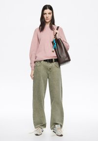 Maglione rosa chiaro lavorato a maglia con maniche lunghe, jeans beige a gamba larga, sneakers grigie e una borsa tote marrone con accenti di portachiavi colorati.
