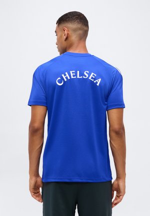 Homme de dos portant un maillot de sport bleu avec "CHELSEA" imprimé en blanc dans le dos sur un fond clair uni.
