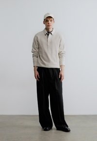 Lysebeige langærmet poloshirt med krave, kombineret med løse sorte bukser og sorte sko. Hvid kasket båret skævt.