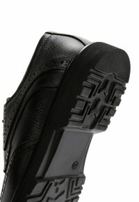 Chaussure en cuir noir avec une surface texturée, dotée d'une semelle en caoutchouc robuste avec un motif de crampons détaillé et des perforations de style brogue.