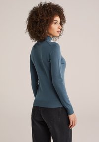 Pull en teal ajusté avec un col haut et des manches longues. Ourlets et poignets côtelés. Texture douce et design épuré, porté avec un jean foncé taille haute.