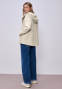 Mujer de pie, vestida con una chaqueta beige con capucha, jeans anchos azules y zapatillas blancas, mirando por encima del hombro.