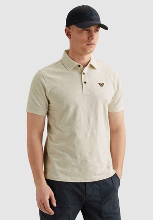 Homme portant un polo beige texturé avec un petit emblème sur la poitrine, casquette noire et pantalon cargo foncé, debout avec une main dans la poche.
