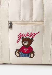 Krämfärgad quiltad väska med en björnmaskot i röd tröja och blå jeans, med "guess" broderat i rött ovanför. Detalj med dragkedjefack.