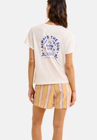 Kremowa bawełniana koszulka z niebieskim nadrukiem tekstowym i wzorem na plecach. Połączona z żółtymi szortami w pionowe niebieskie paski.
