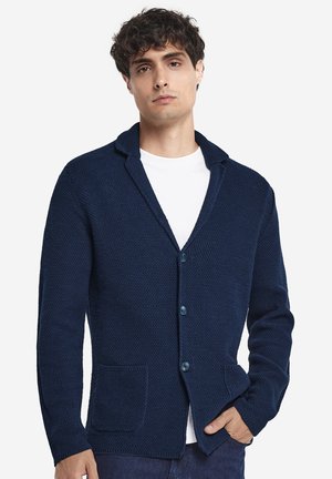 Jeune homme portant un cardigan en tricot texturé bleu marine avec des boutons sur un t-shirt blanc, posant avec une main dans la poche.