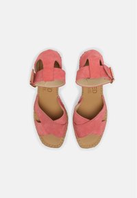 Un paio di sandali di suede rosa con fasce incrociate sopra le dita, cinturini alla caviglia con fibbie e suole cucite color tan su sfondo bianco.